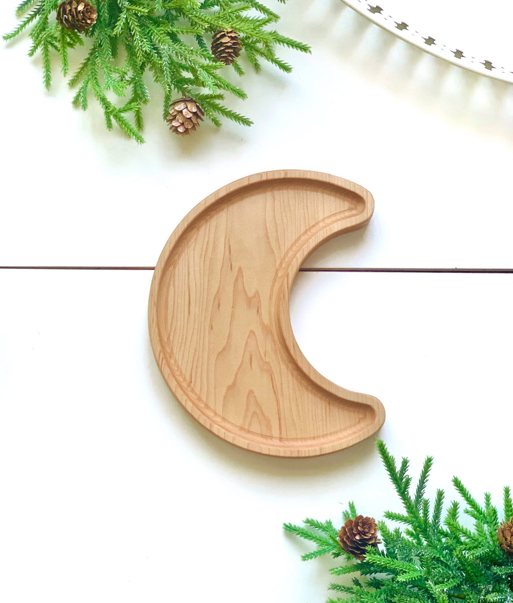 Natural wooden crescent moon shaped sensory tray surrounded by green pine branches and pinecones on white surface