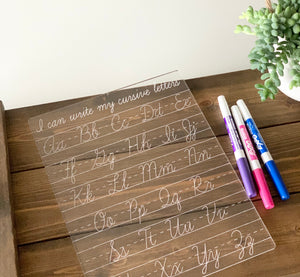 Clear acrylic dry erase board engraved with cursive alphabet letters and three colorful markers on wooden surface