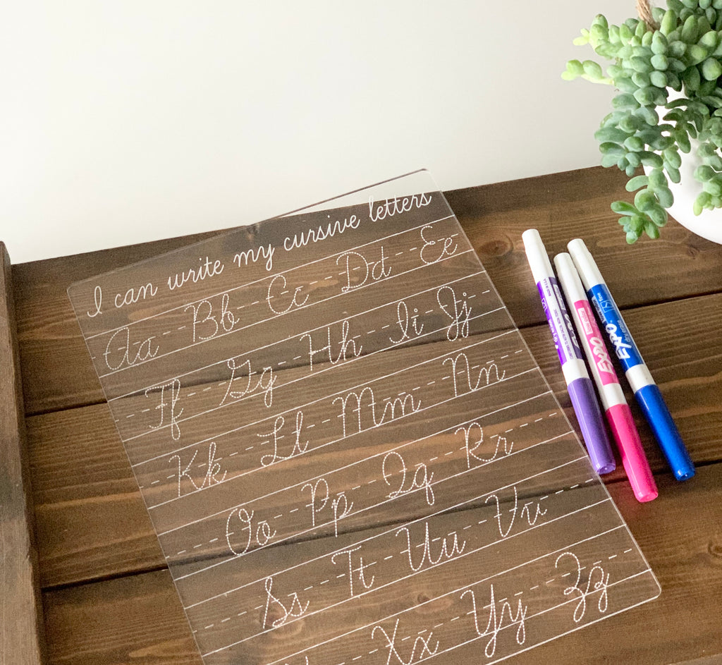 Clear acrylic dry erase board engraved with cursive alphabet letters and three colorful markers on wooden surface