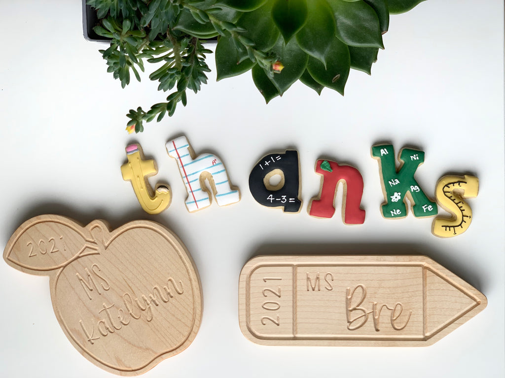 Colorful alphabet magnets spelling TEACHERS with engraved wooden apple and pencil name plaques on white surface with green plant