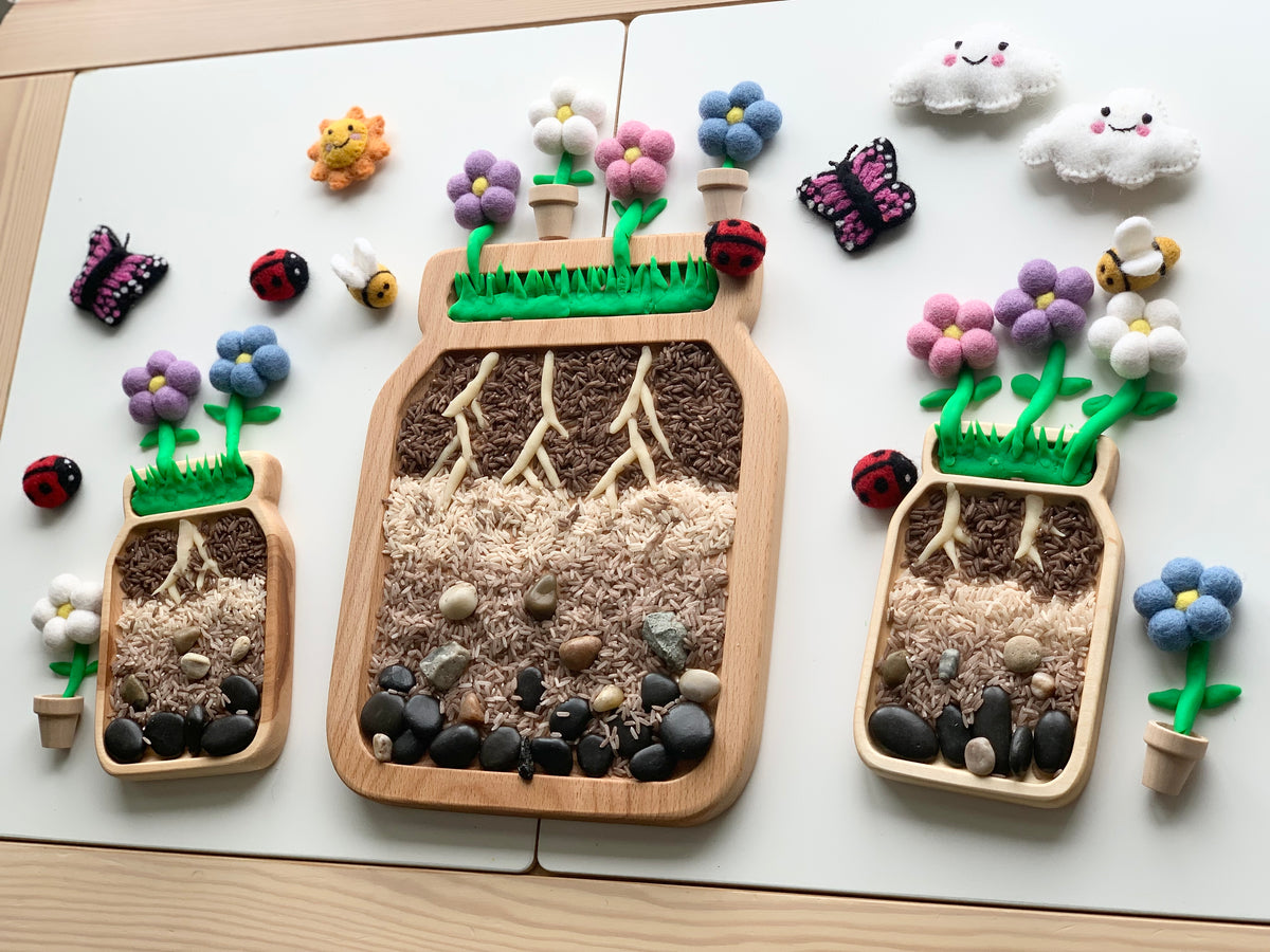 Three large wooden jar plate sensory trays filled with natural materials and decorated with felt flowers and insects on white surface