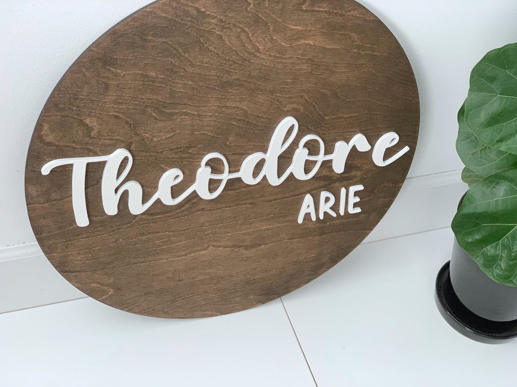 Round wooden 3D name sign stained brown with white cursive and block letters on white floor near green plant