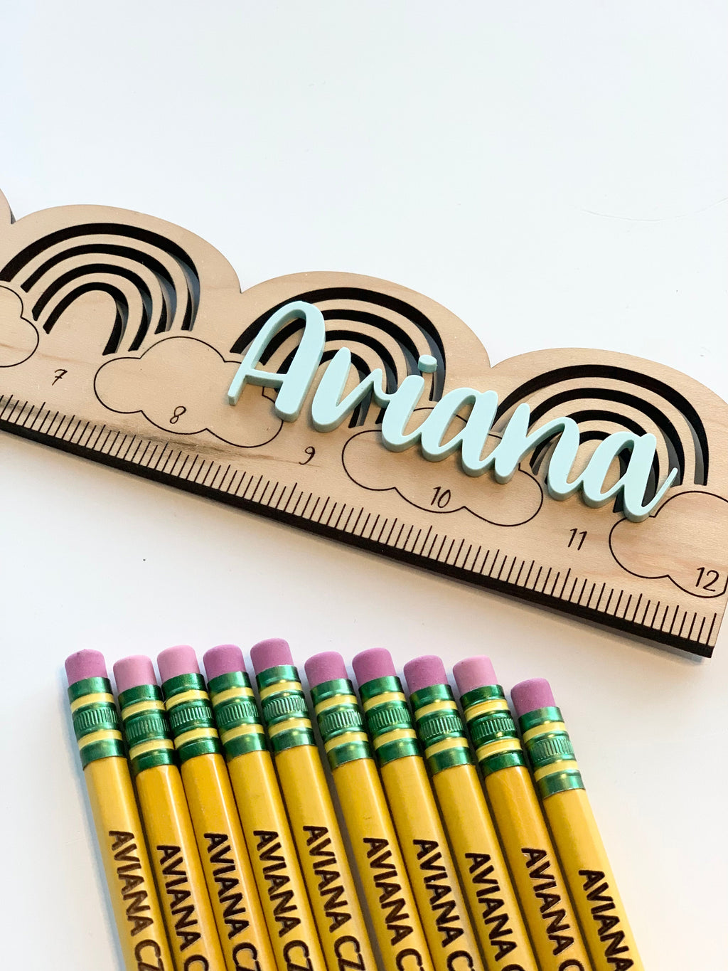 Yellow personalized pencils with green ferrules and pink erasers next to a wooden rainbow ruler with the name Ariana in blue cursive letters