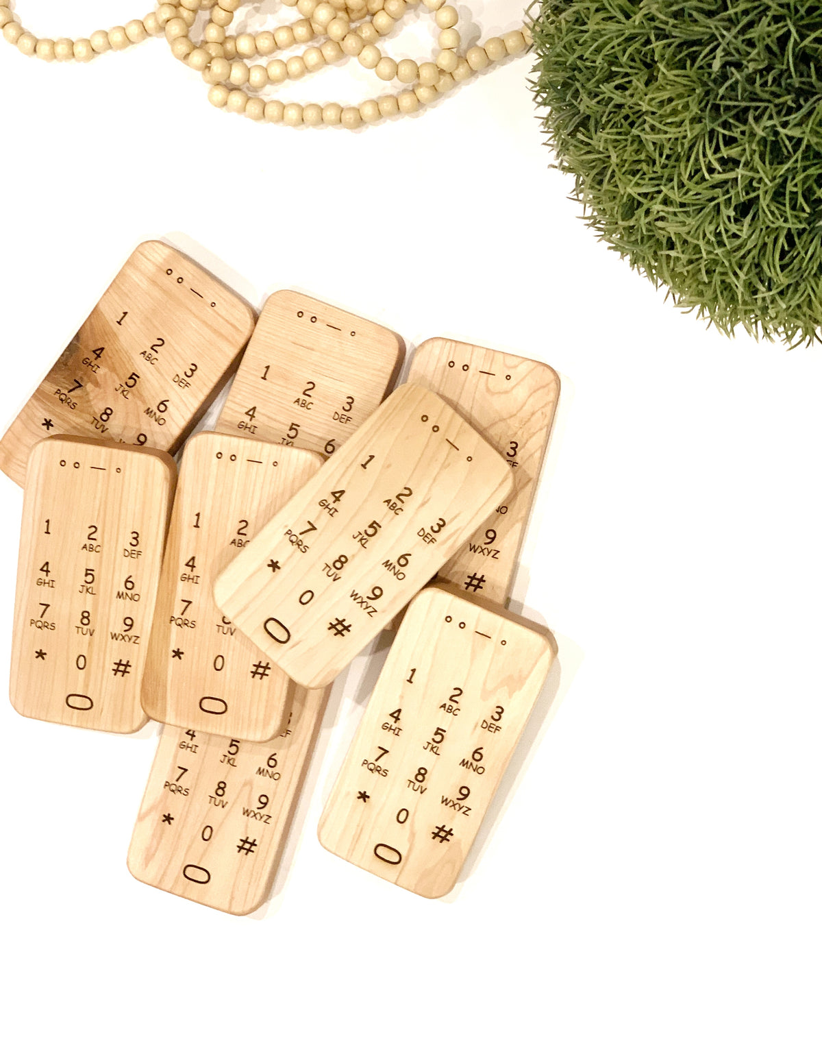 Set of natural wooden toy phones with engraved numbers and symbols on a white surface with greenery and beads nearby