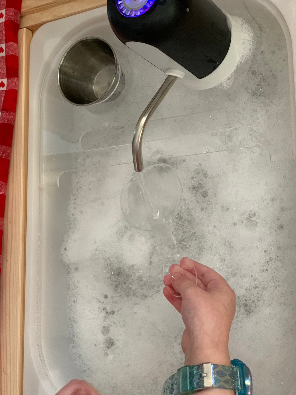 White play kitchen sink filled with bubbles and water pouring from black faucet into clear cup held by child’s hands