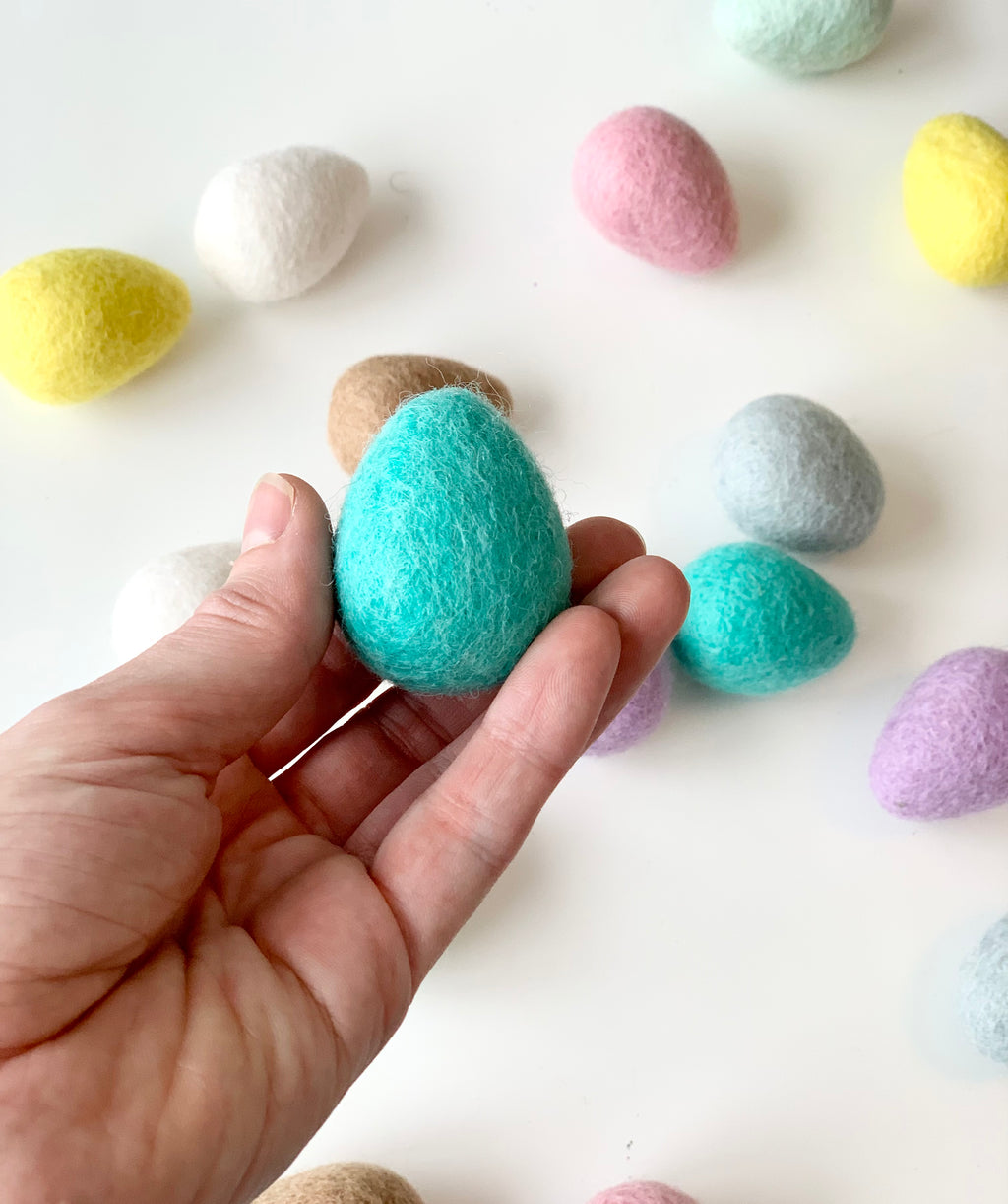 Hand holding one turquoise felt Easter egg with additional pastel-colored felt eggs scattered on white surface