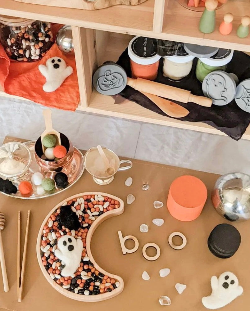 Moon-shaped wooden sensory tray filled with beans and felt ghost figures on a brown table surface
