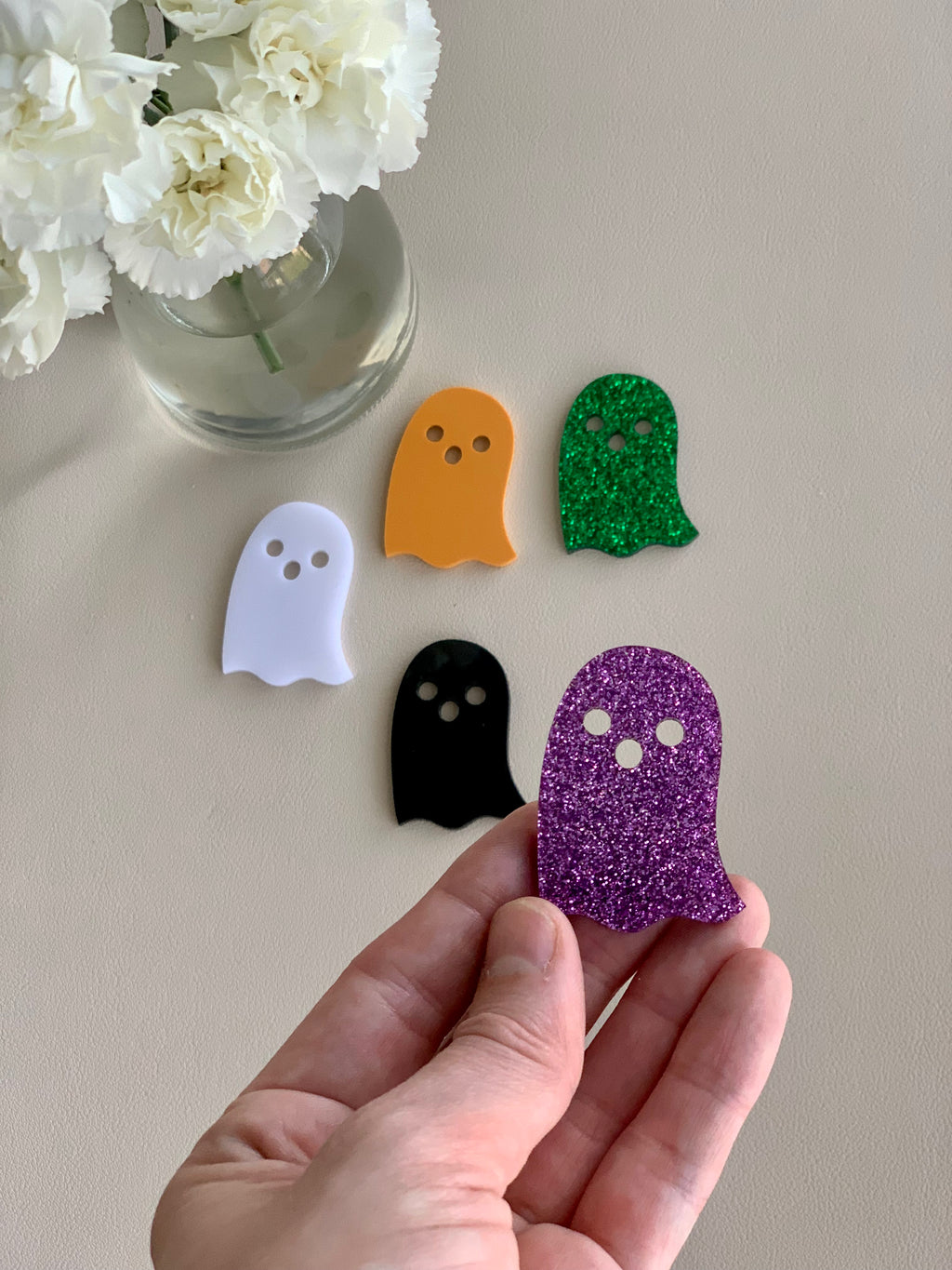 Set of five colorful acrylic ghost shapes in white, orange, black, green glitter, and purple glitter on beige surface
