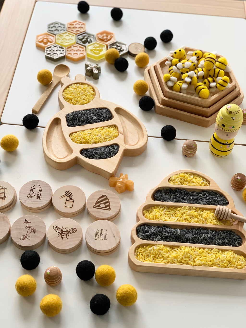 Wooden bee-shaped sensory tray and hive tray filled with yellow and black rice alongside wooden discs and bee toys on white surface