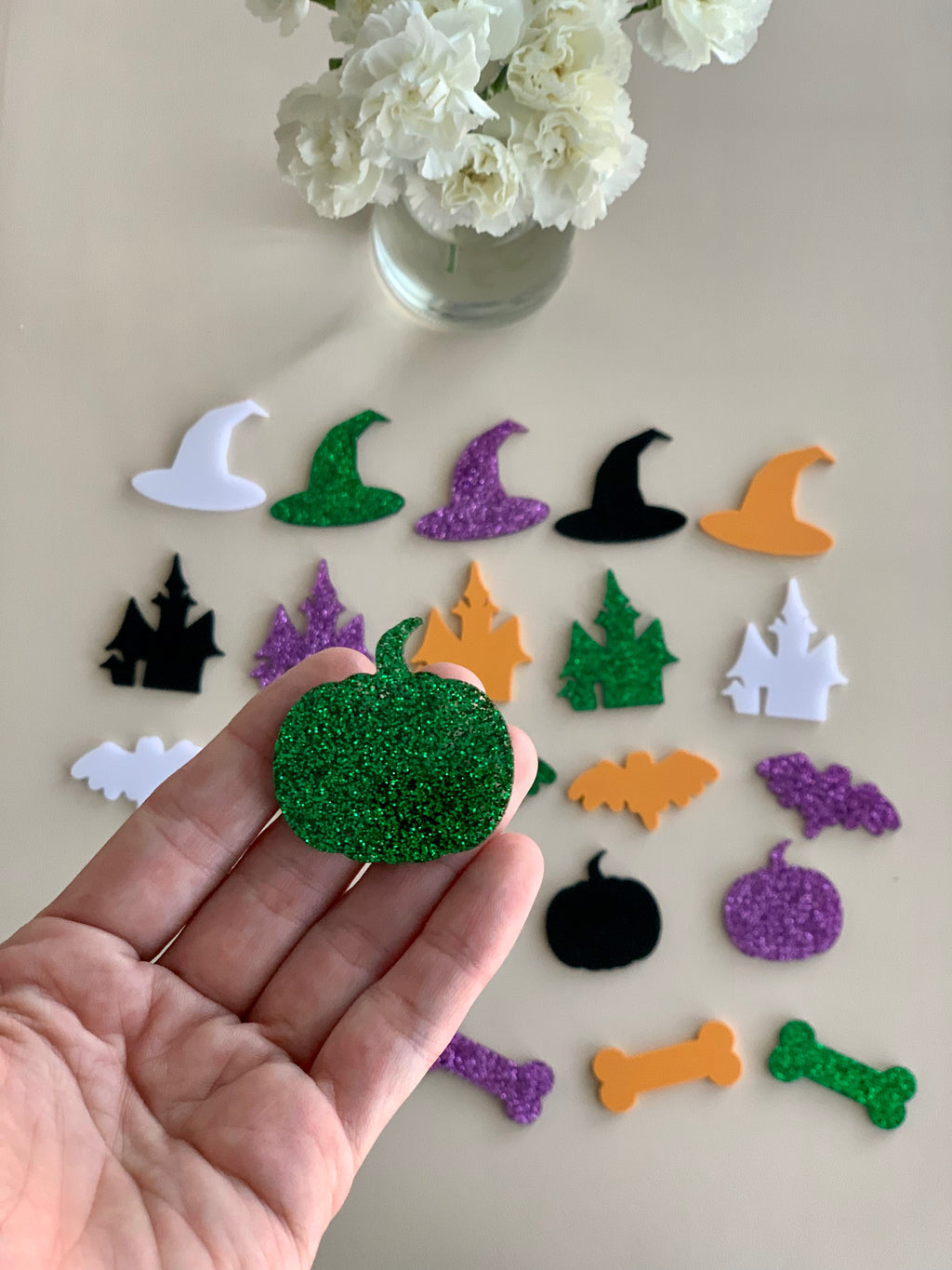 Hand holding glittery green pumpkin acrylic shape with various Halloween-themed loose parts on beige surface and flowers above