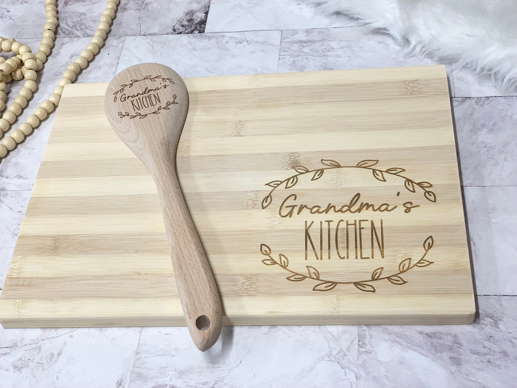 Personalized wooden spoon and cutting board set engraved with "Grandma's Kitchen" and decorative leaf design on light wood background