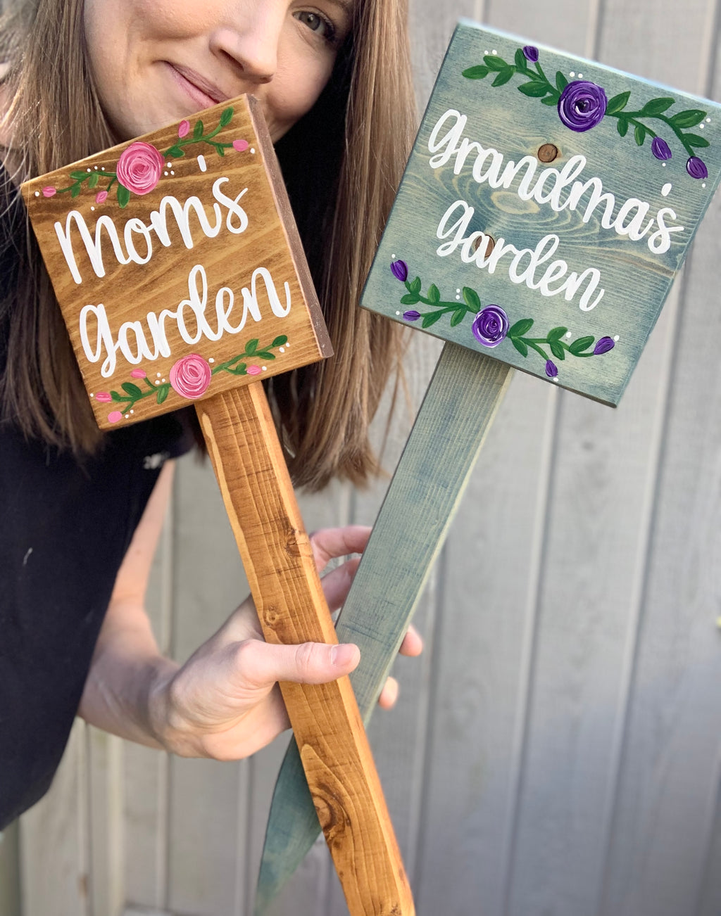 Two small square wooden garden signs with floral decorations reading Moms garden and Grandmas garden held by person in black shirt