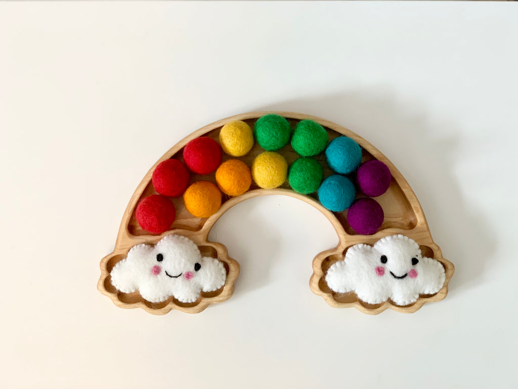 Wooden rainbow sensory tray with colorful felt balls arranged in arch and two smiling felt clouds on white background