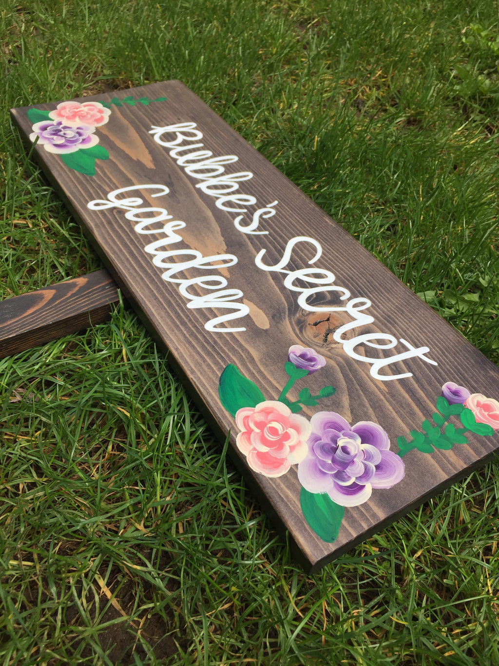 Dark stained custom wooden garden sign with white cursive letters and hand-painted pink and purple flowers on grass background