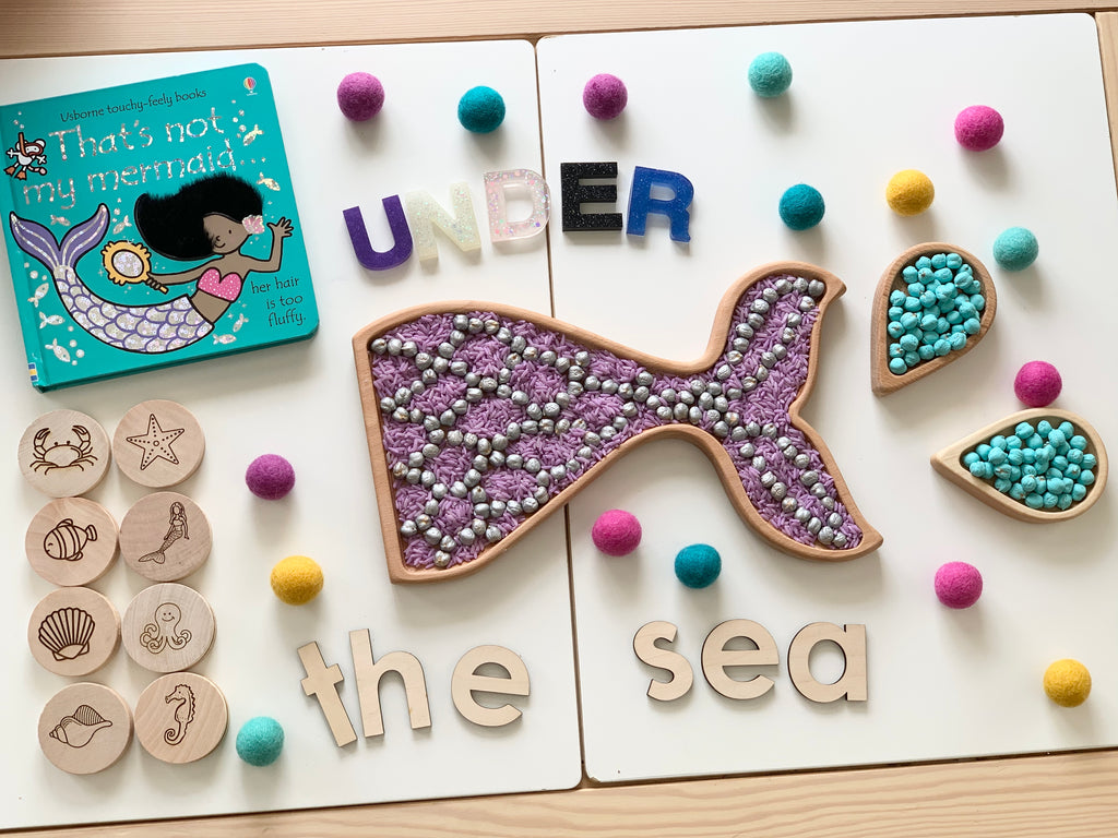 Mermaid tail sensory tray with purple beads and wooden sea creature tokens arranged on white background with book and colorful felt balls