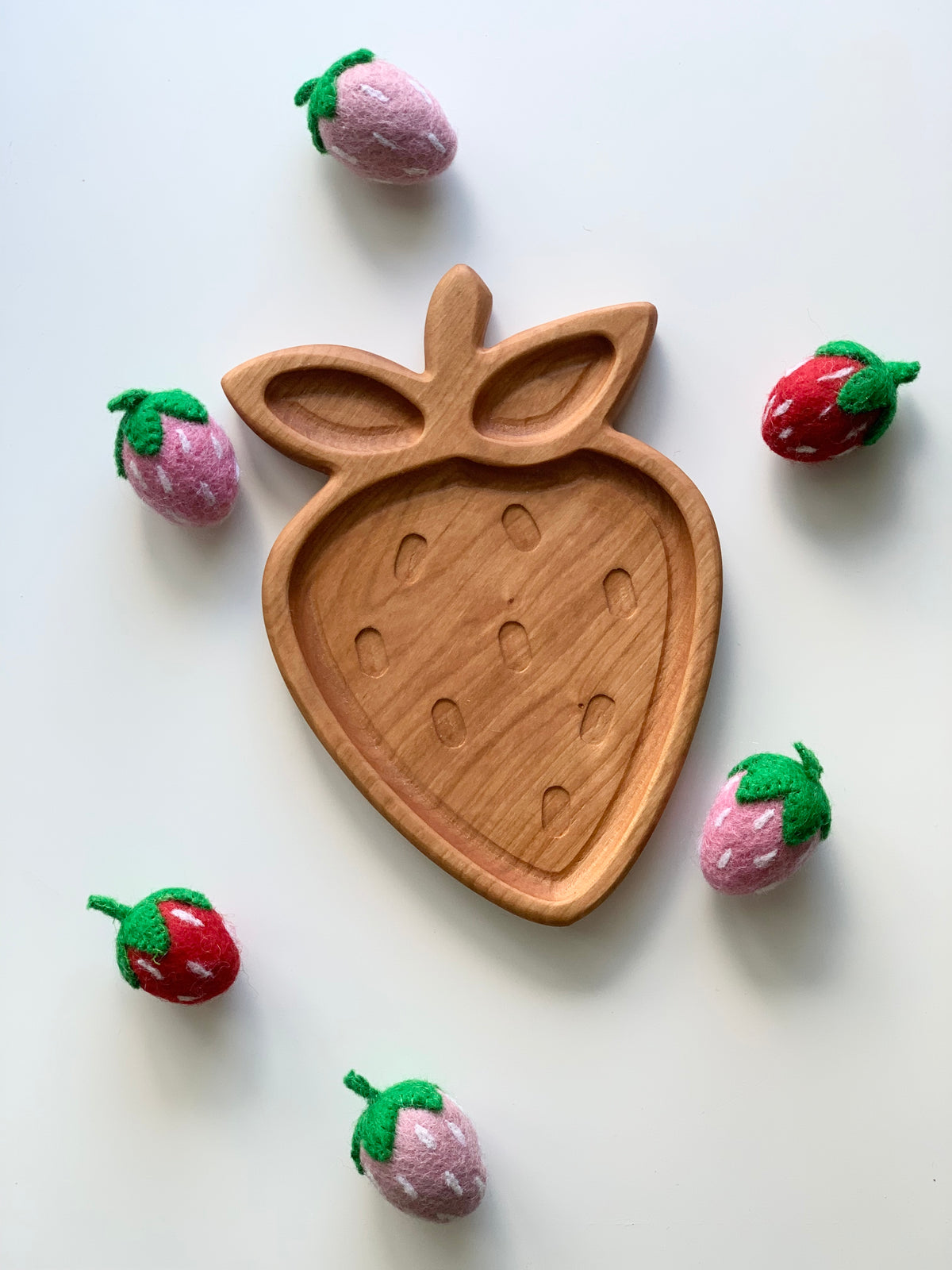 Cherry wood strawberry-shaped sensory tray surrounded by small red and pink felt strawberries on white background
