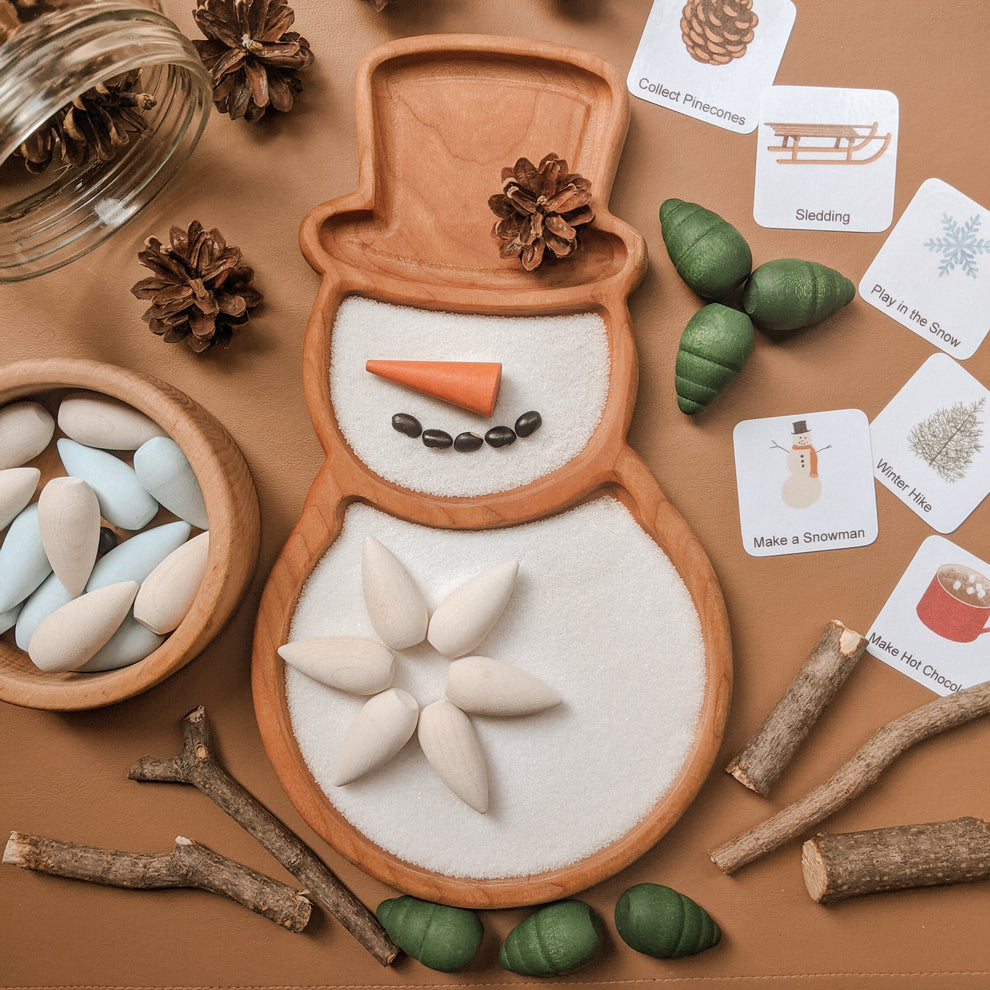 Snow person sensory tray with wooden carrot nose, coffee bean smile, and white wooden pieces on brown background with nature elements