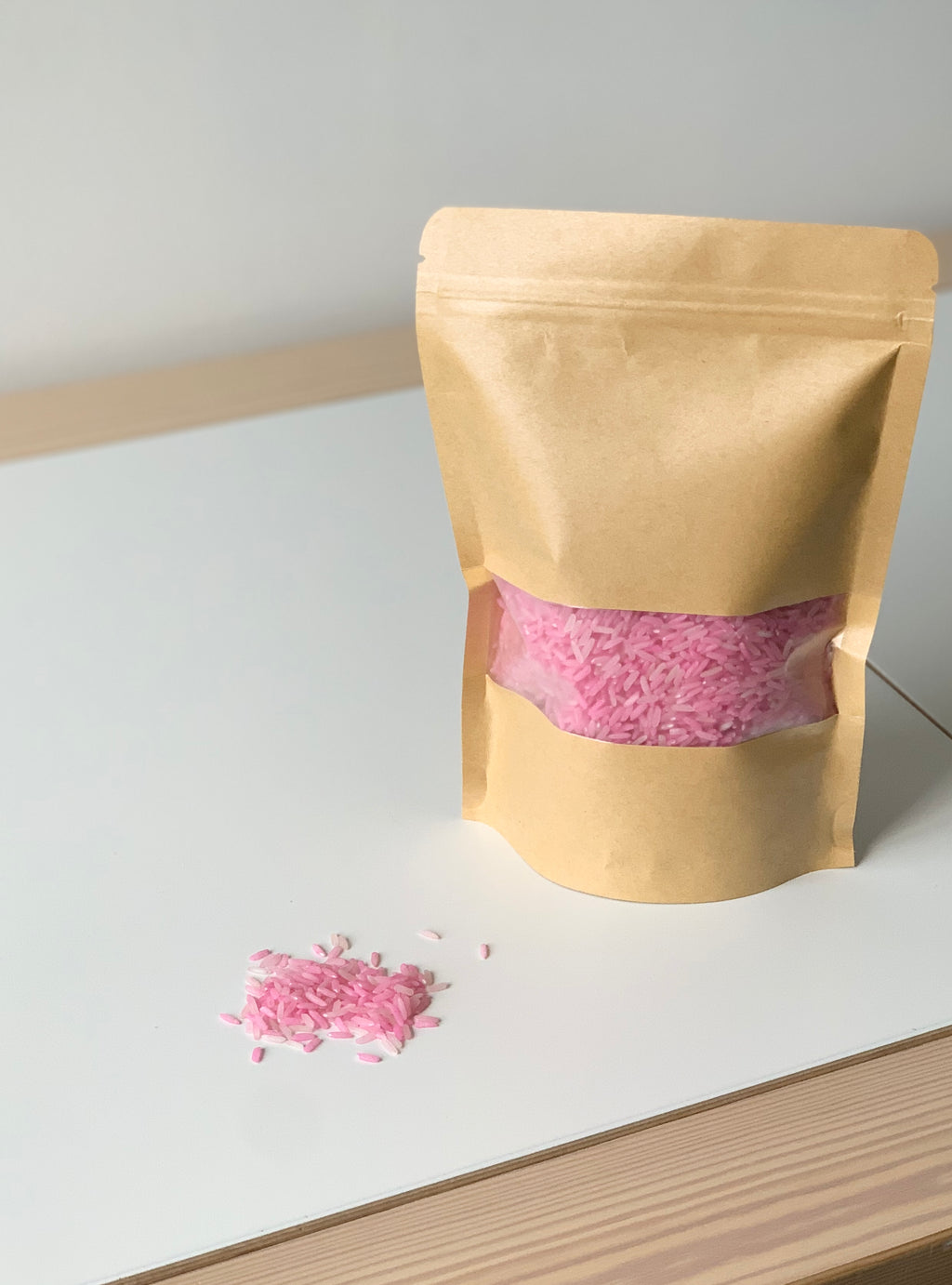 Kraft paper pouch with transparent window showing pink sensory rice filler spilled on white and wood surface