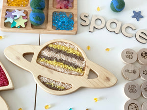 Natural wooden sensory tray shaped like the planet Saturn filled with colored rice and surrounded by wooden space-themed toys