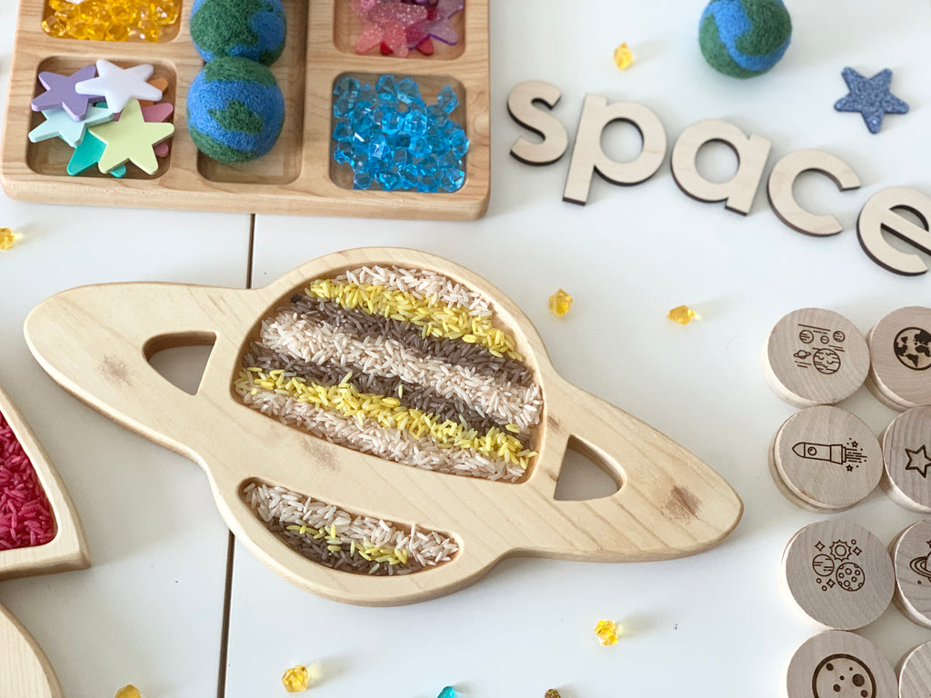 Natural wooden sensory tray shaped like the planet Saturn filled with colored rice and surrounded by wooden space-themed toys