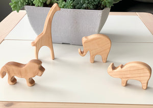 Natural wooden safari animal toy figurines including giraffe, elephant, lion, and rhinoceros on white surface with plant background