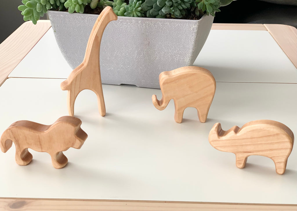 Natural wooden safari animal toy figurines including giraffe, elephant, lion, and rhinoceros on white surface with plant background
