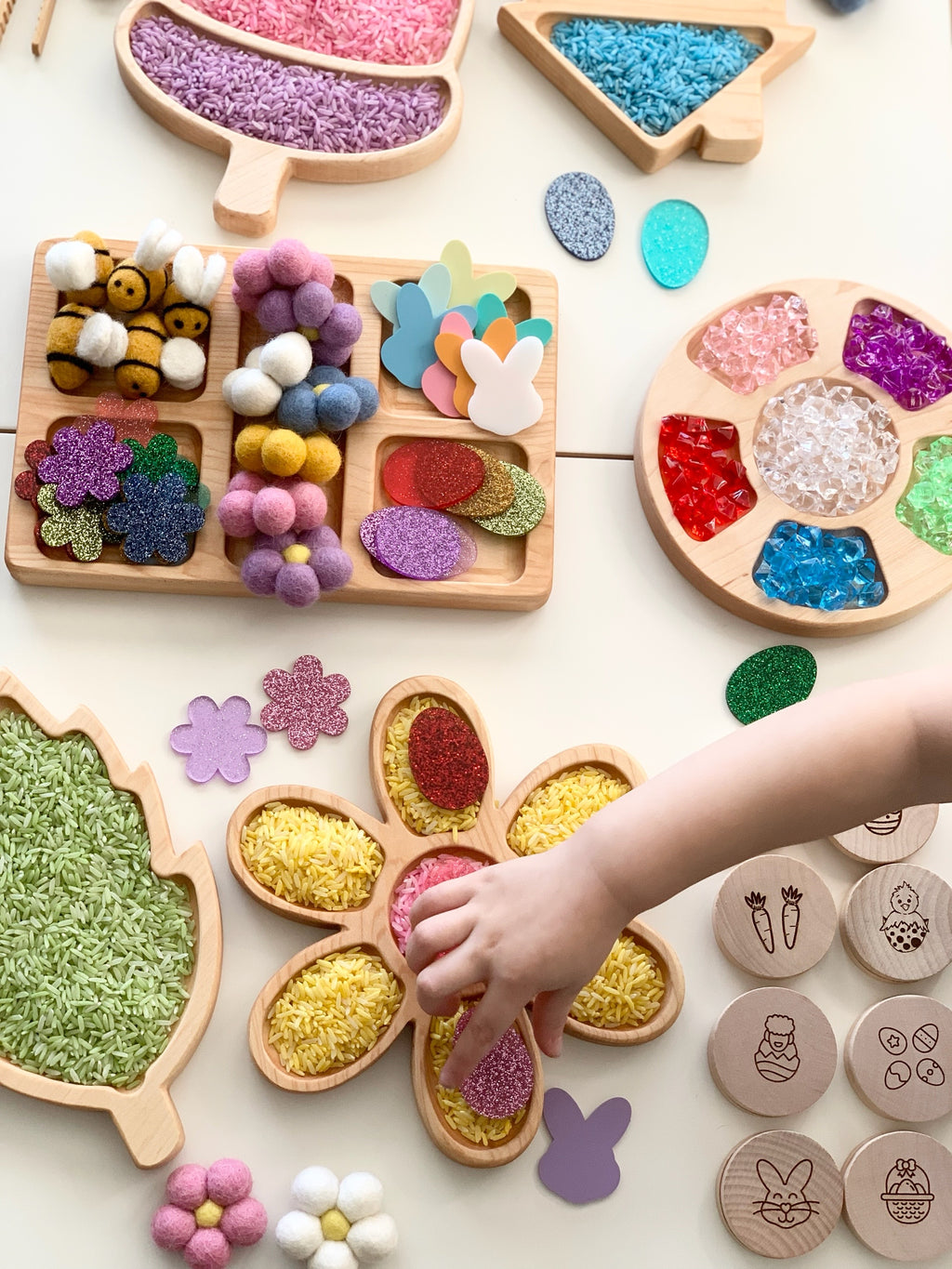 Natural wooden rectangular tinker tray filled with colorful felt balls, glitter shapes, and paper cutouts on white surface