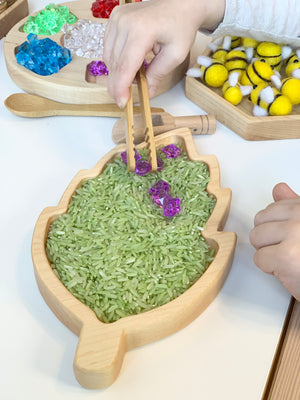 Birch leaf shaped wooden sensory tray filled with green rice and child using wooden tongs to pick purple gems