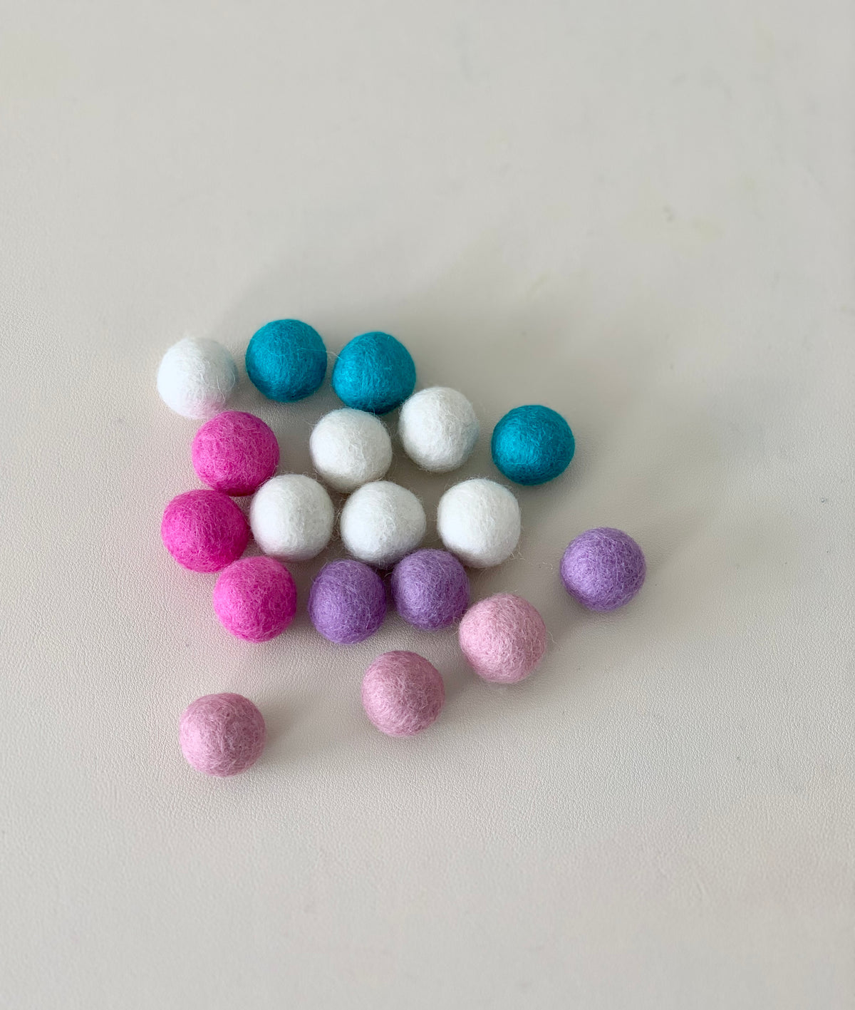 Assorted pastel felt balls in pink, purple, white, and teal shades arranged on a white surface