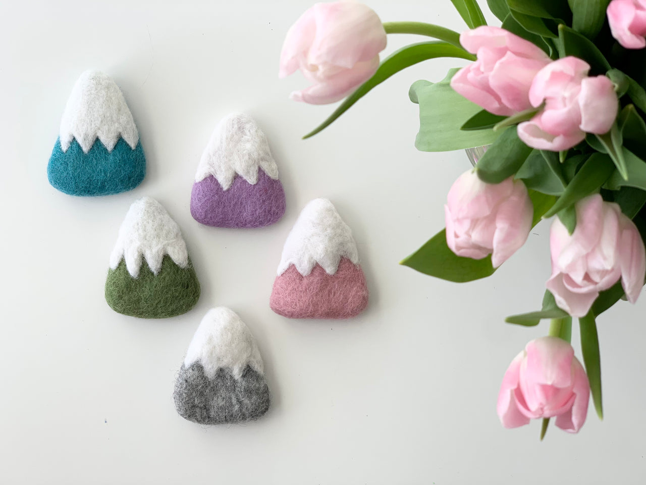 Five handmade felt mountains in blue, purple, green, pink, and gray with white snowy tops on white background with pink tulips