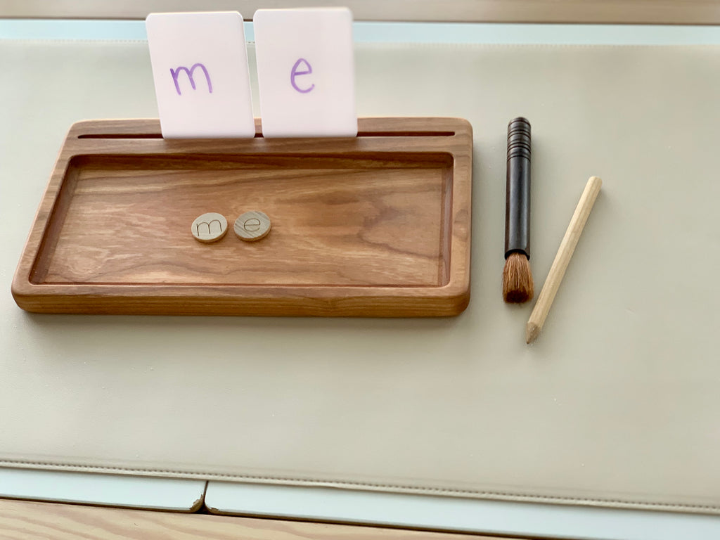Rectangle wooden sensory tray with flash cards, letter tiles spelling "me", small brush, and wooden stylus on white surface