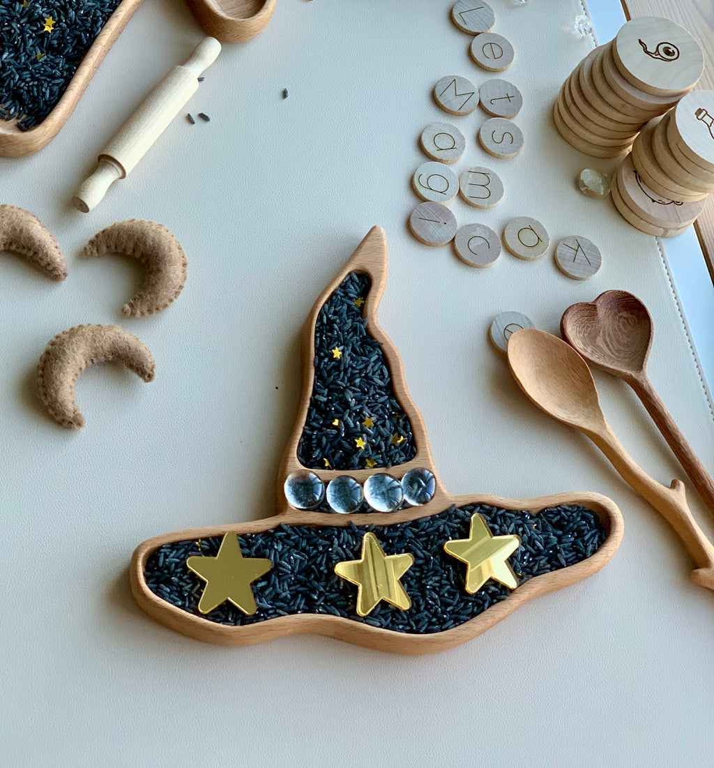 Wooden witch hat sensory tray filled with black rice and decorated with gold stars and clear stones on white surface