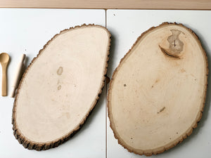 Two large live edge wooden trays with smooth surfaces and natural bark edges on white background with wooden utensils nearby