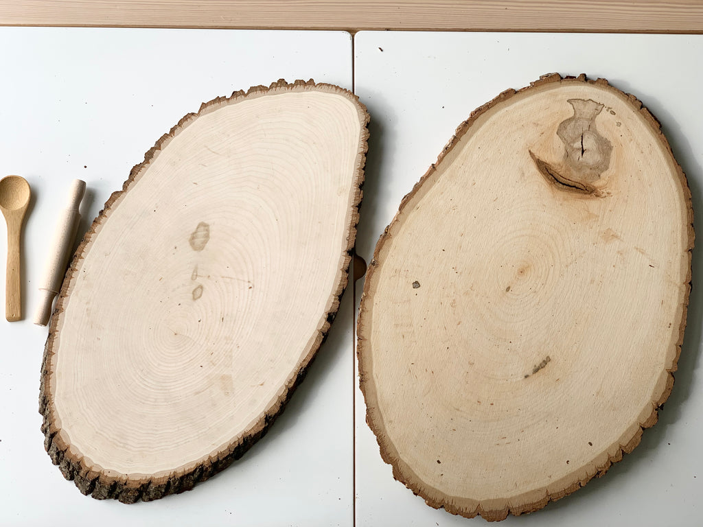 Two large live edge wooden trays with smooth surfaces and natural bark edges on white background with wooden utensils nearby