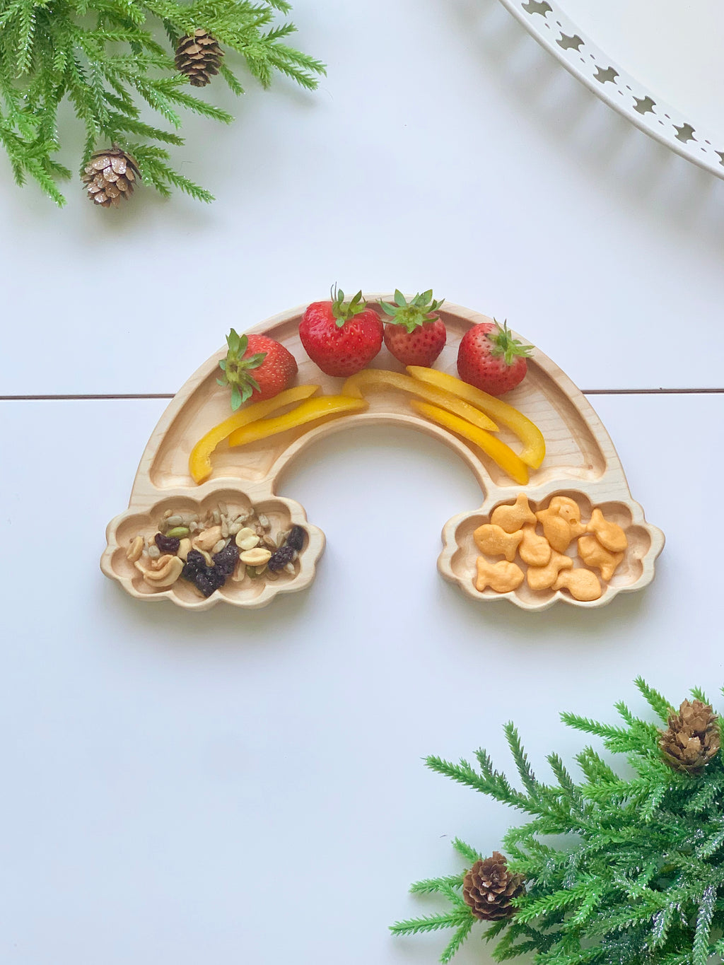 Natural wooden rainbow-shaped sensory tray with sections holding strawberries, yellow pepper strips, nuts, and goldfish crackers on white surface