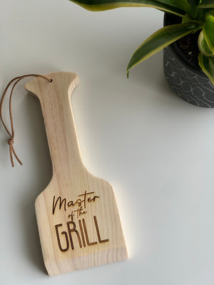 Natural wooden barbecue scraper with leather loop and "Master of the Grill" engraved text on white surface near plant
