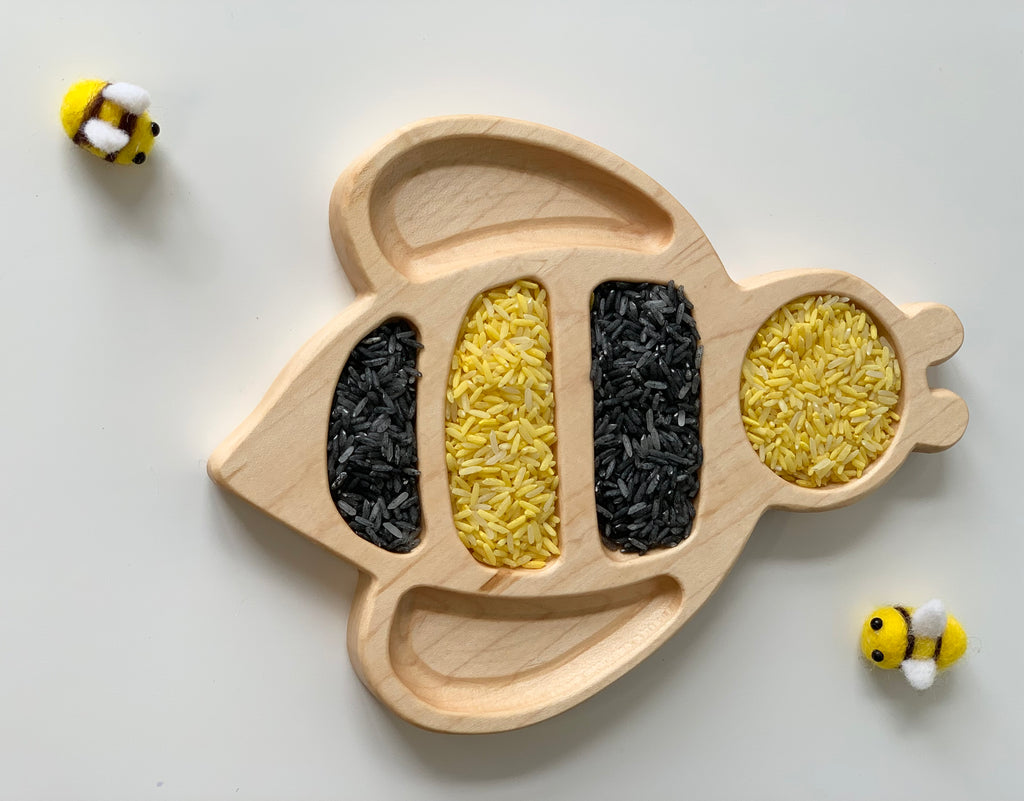 Natural wooden bumble bee shaped sensory tray filled with yellow and black rice with small bee toys on white background
