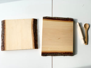 Two clear coated live edge wooden trays with bark edges alongside a wooden rolling pin and spoon on white surface