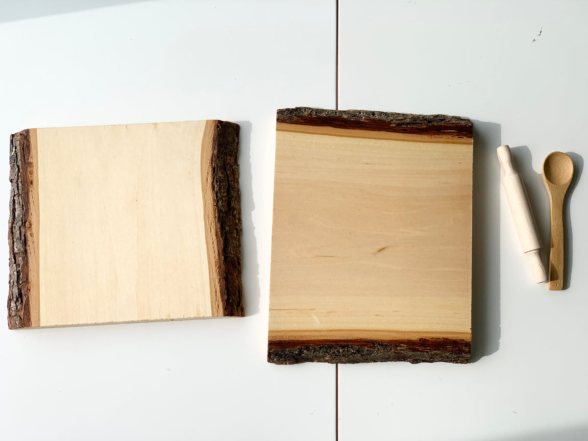 Two clear coated live edge wooden trays with bark edges alongside a wooden rolling pin and spoon on white surface