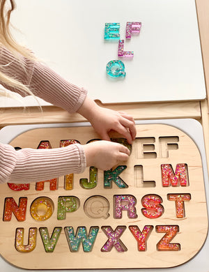 Child’s hands placing colorful resin uppercase letter M into wooden puzzle board with other letters and loose pieces above on white surface