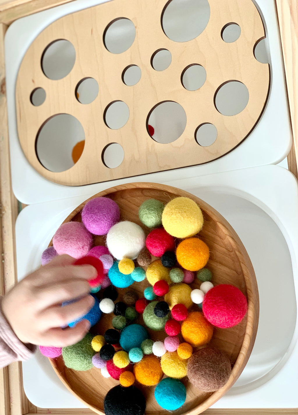 Wooden circle sorter insert with various-sized colorful felt balls and a child’s hand on white surface