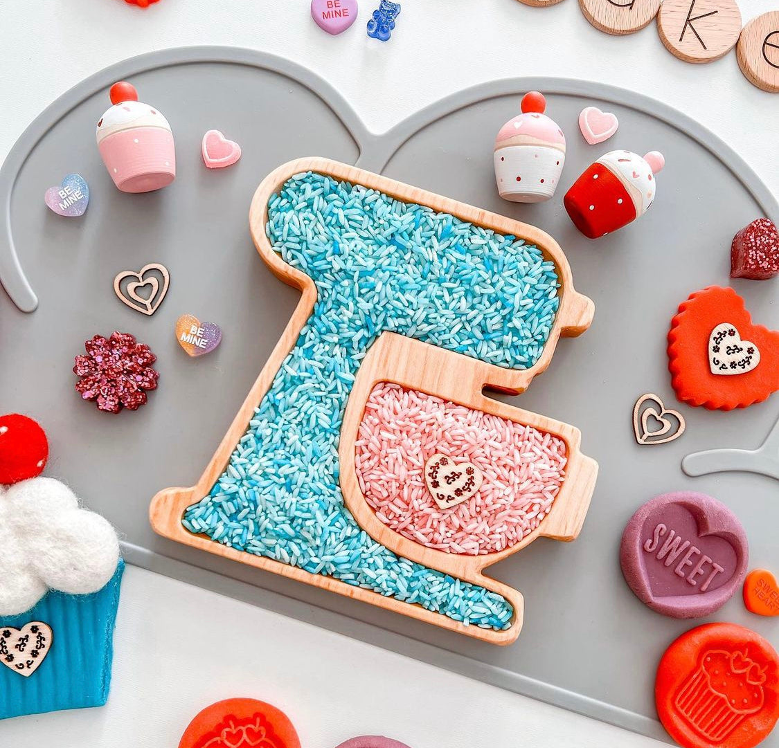Wooden mixer plate sensory tray filled with blue and pink rice on gray heart-shaped tray with small candy decorations
