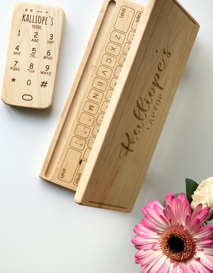 Personalized wooden toy phone and keyboard with engraved letters and names on natural wood, flat on white surface with pink flower