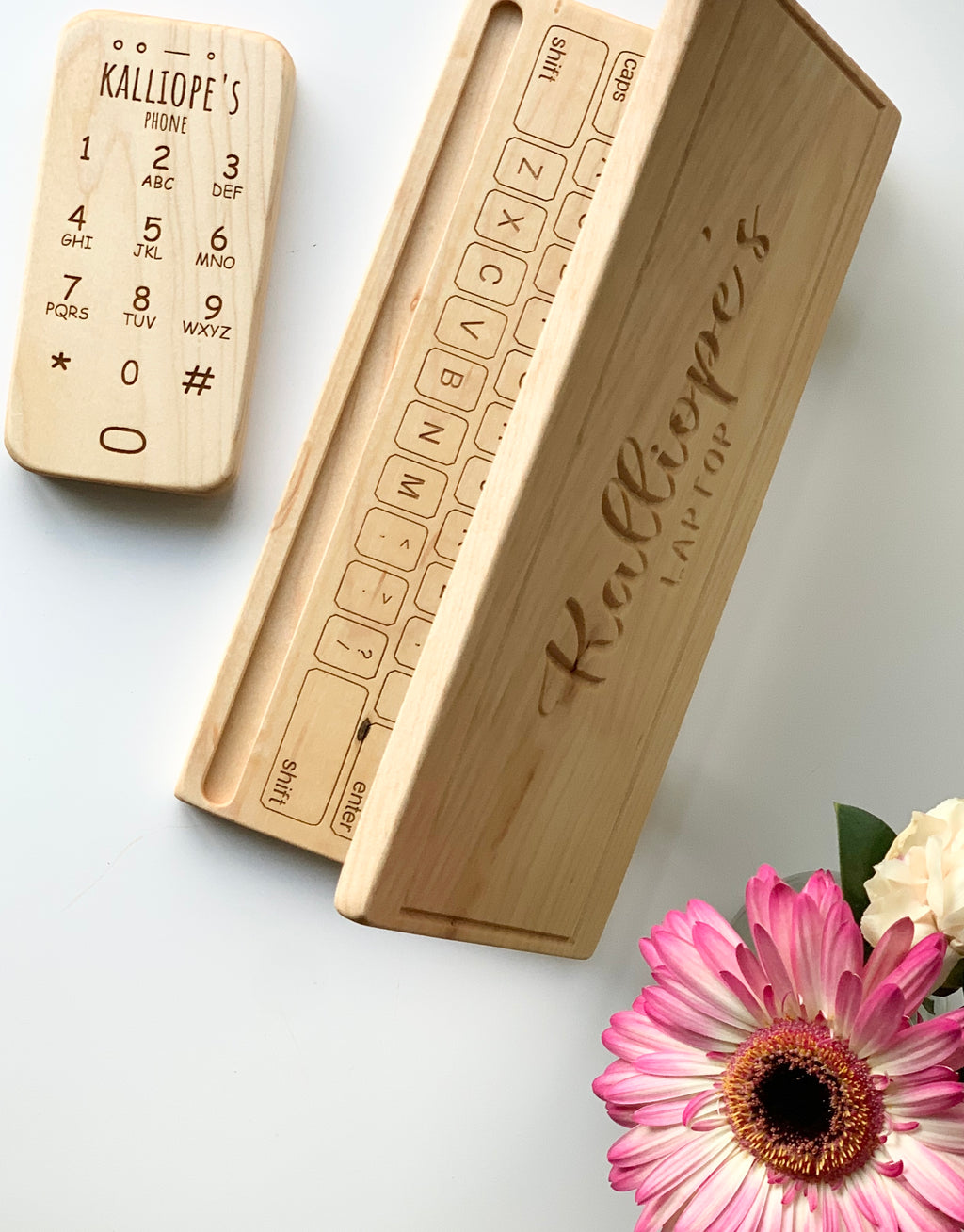 Personalized wooden toy phone and keyboard with engraved letters and names on natural wood, flat on white surface with pink flower