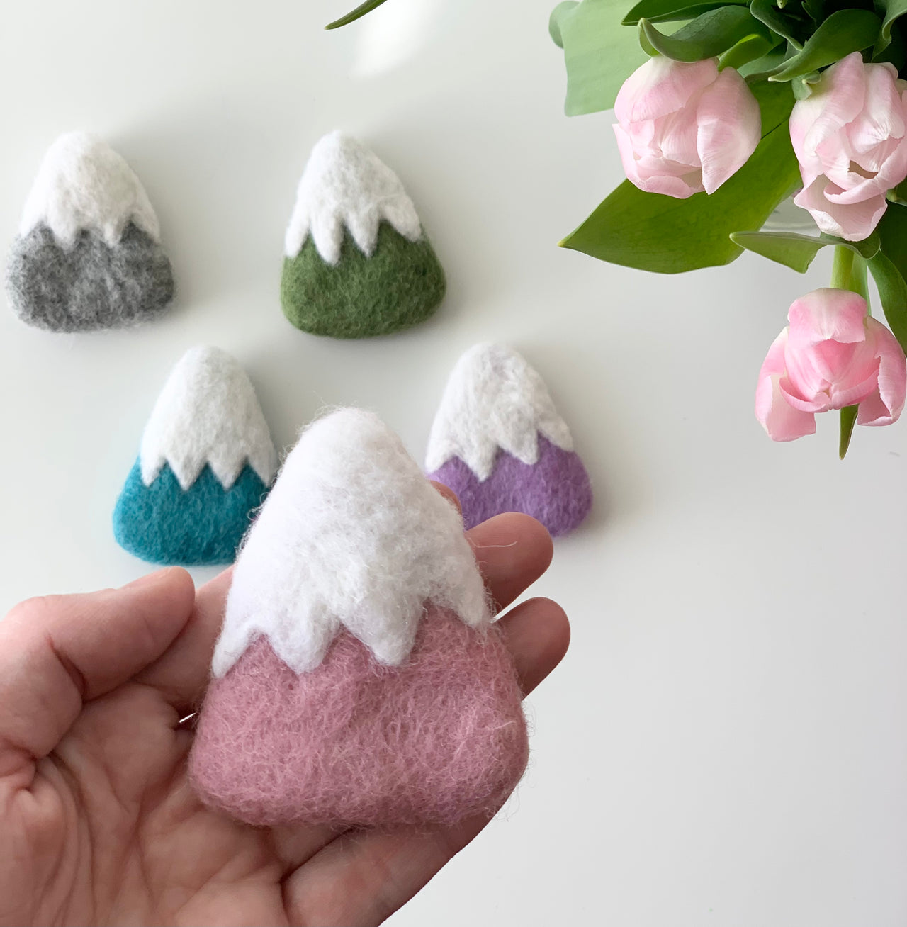 Hand holding one pink felt mountain with white snowcap with four other colorful felt mountains and pink flowers on white surface