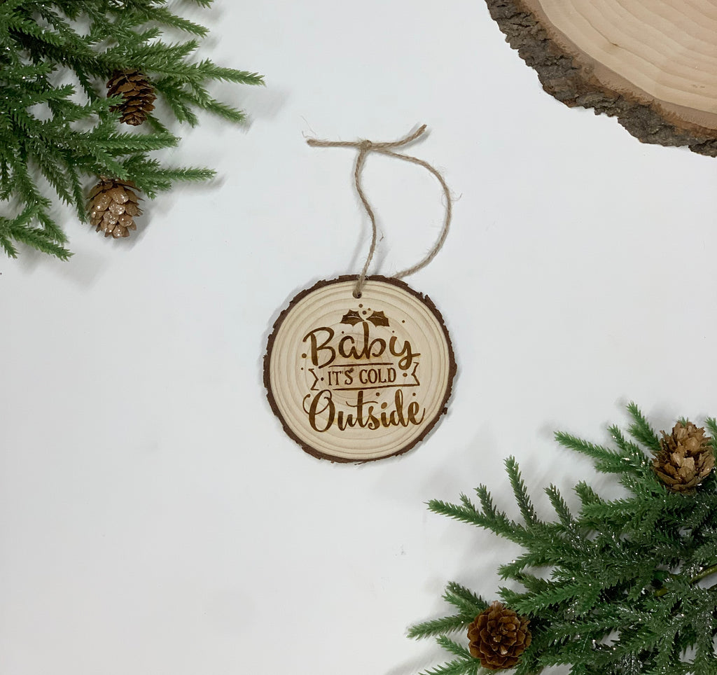 Round wooden Christmas ornament with wood burned "Baby it’s Cold Outside" text and antler graphic hanging on twine string