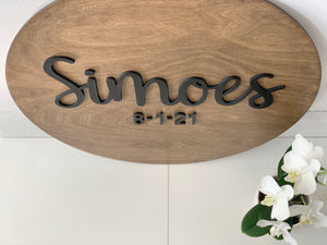 Oval wooden sign with black 3D cursive last name and date letters on natural wood background near white orchids