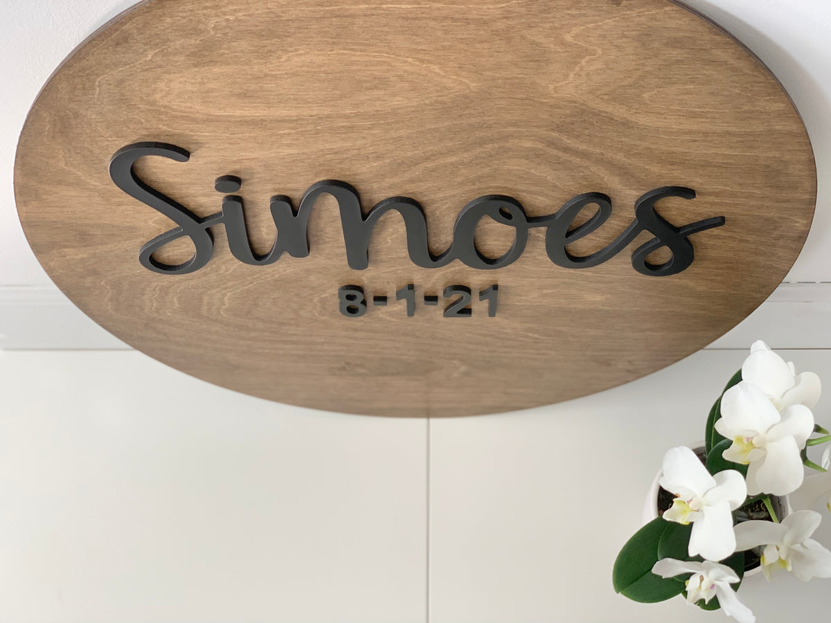 Oval wooden sign with black 3D cursive last name and date letters on natural wood background near white orchids