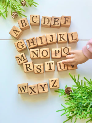 Natural 1.5-inch wooden alphabet blocks with engraved black uppercase letters arranged on white surface with greenery and child’s hand