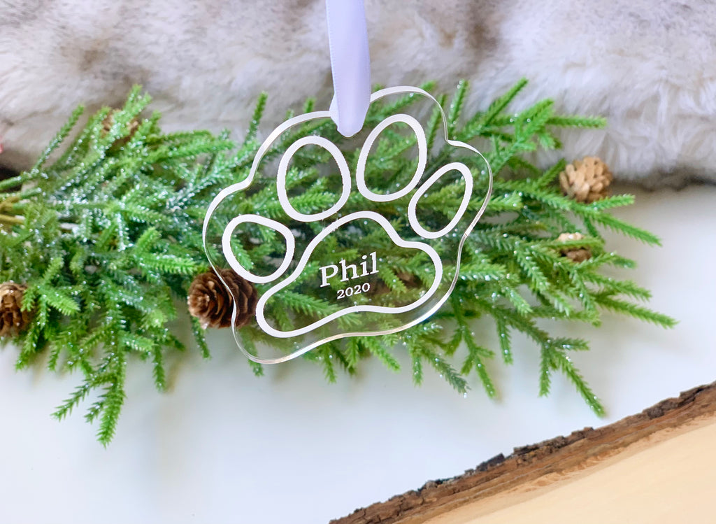 Clear acrylic paw print pet memorial Christmas ornament engraved with name and year hanging on greenery background